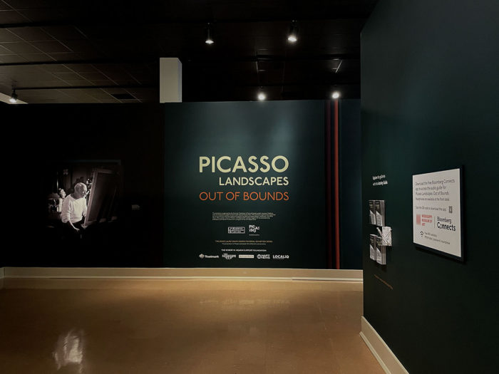Entry area to Picasso Landscapes exhibition. The title wall is centered, there is a life-sized photograph of Picasso on the left and area on right for picking up gallery guides and learning how to access the museum's app.